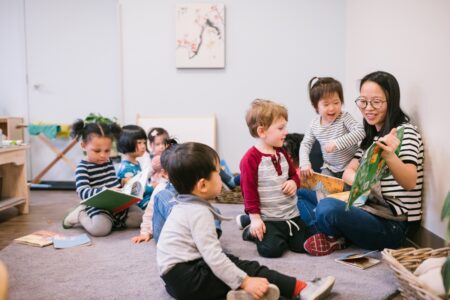 a teacher sitting on a classroom floor surrounded by kindergarten-aged children.