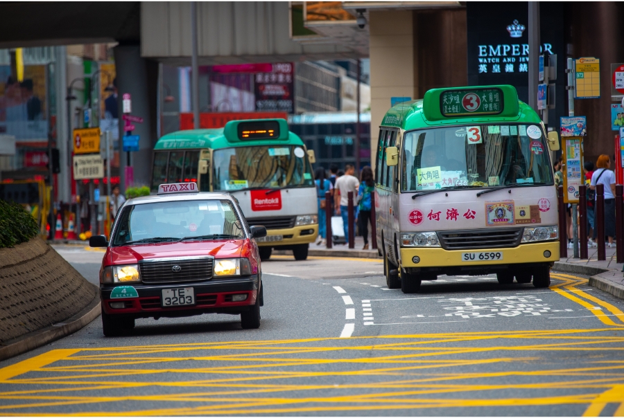 A red Hong Kong taxi drives down a road followed by two yellow-and-green light buses.