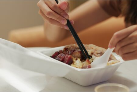 A person eats Hong Kong char siu out of a disposable box using chopsticks and a plastic spoon.