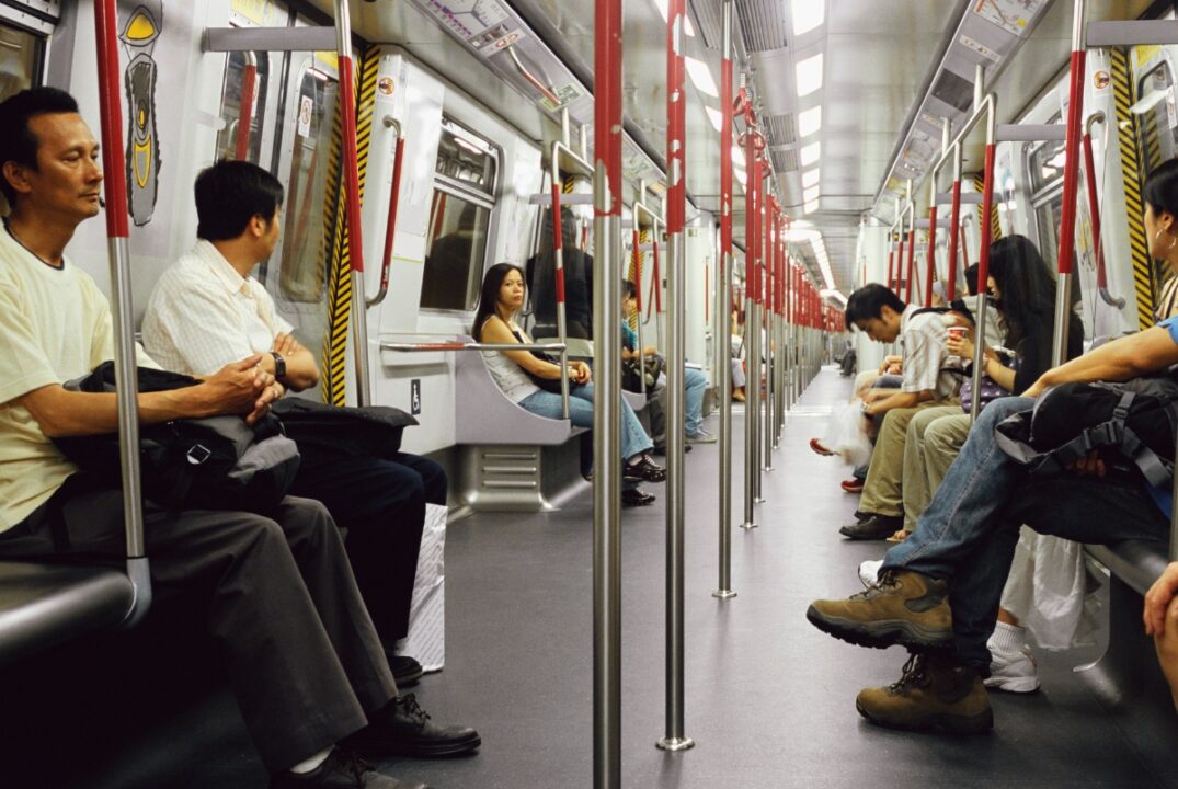 People sit in a compartment of a Hong Kong MTR subway train. The sides of the train are lined with seats upon which passengers sit, and there are poles running down the middle of the train.