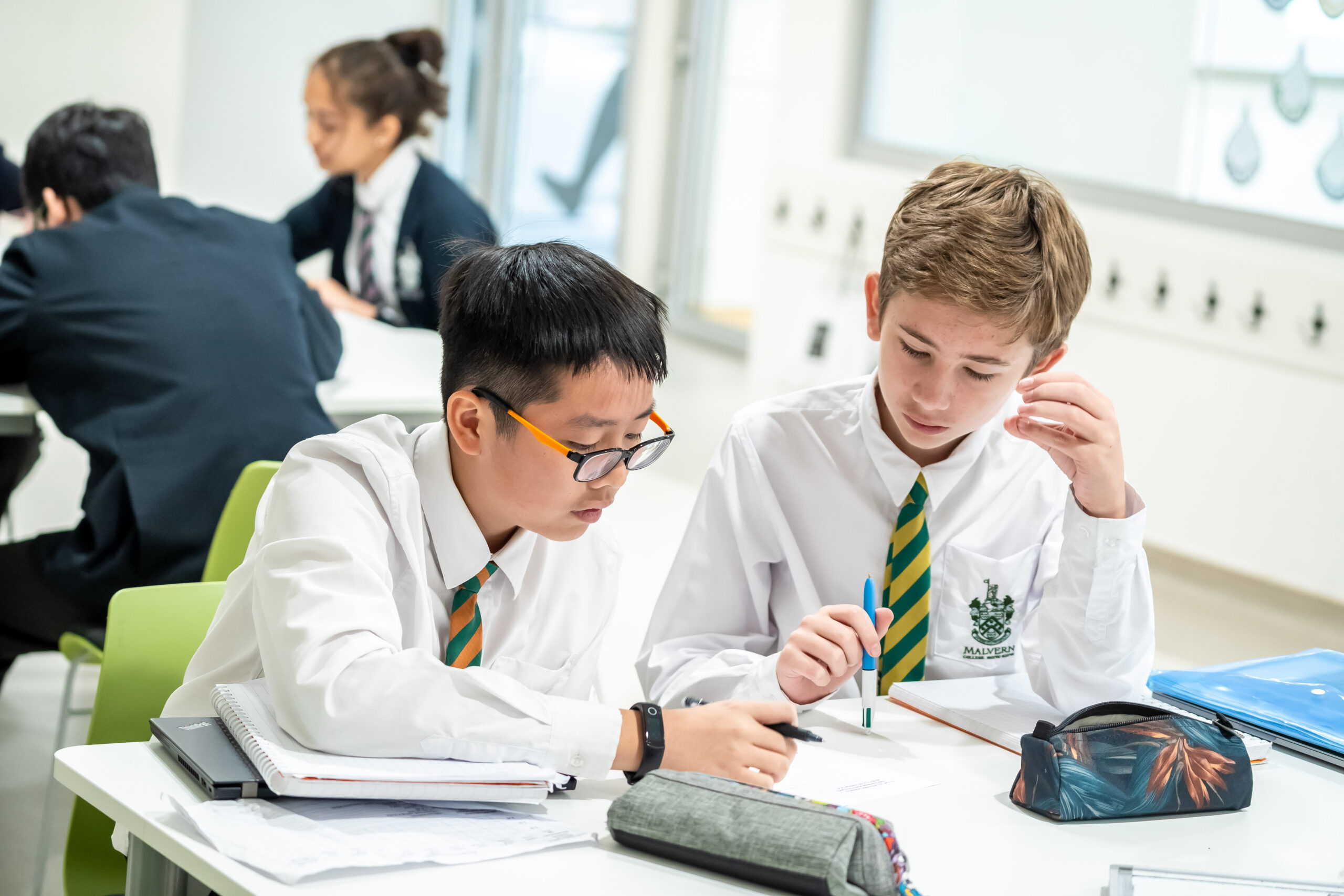 Students work on their assignments at Malvern College Hong Kong. The two students in focus are boys wearing the school's uniform -- white collared long-sleeved shirts with striped ties. They sit on chairs in front of a white desk with books, a laptop, a folder, and pencil cases on it. They are holding pens while working on the assignment. Other students in the background wear the school blazer over their uniforms while seated at another desk.